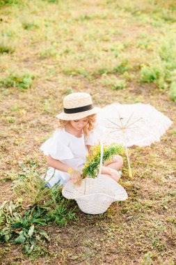 Işık ile tatlı bir küçük kız bir hasır şapka, beyaz bir elbise ve dantel şemsiye yaz tarlada çiçekler bir sepet ile bukleler. Yaz aylarında kırsal yaşam
