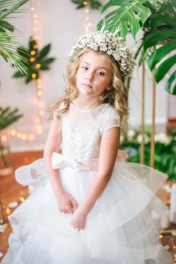 Beyaz gelinlik ve çelenk çiçek süslemeleri yapılmış tropik yapraklar, beyaz güller ve orkide çiçek dolu sarışın kıvırcık saçlı sevimli küçük kız