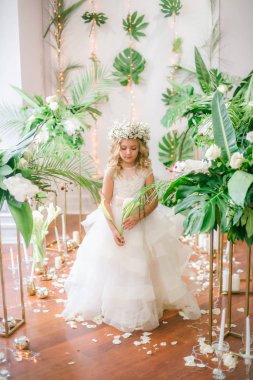 Beyaz gelinlik ve çelenk çiçek süslemeleri yapılmış tropik yapraklar, beyaz güller ve orkide çiçek dolu sarışın kıvırcık saçlı sevimli küçük kız