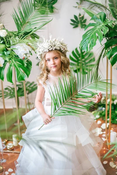 Beyaz gelinlik ve çelenk çiçek süslemeleri yapılmış tropik yapraklar, beyaz güller ve orkide çiçek dolu sarışın kıvırcık saçlı sevimli küçük kız