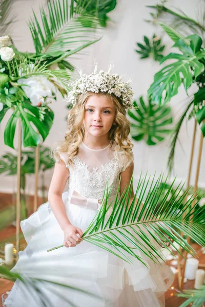 Beyaz gelinlik ve çelenk çiçek süslemeleri yapılmış tropik yapraklar, beyaz güller ve orkide çiçek dolu sarışın kıvırcık saçlı sevimli küçük kız