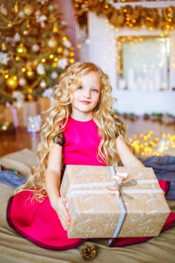 Uzun kıvırcık saçlı sarışın evde hediyeler ve çelenk ve dekore edilmiş bir şömine ile Noel ağacı yakınındaki sevimli küçük kız 
