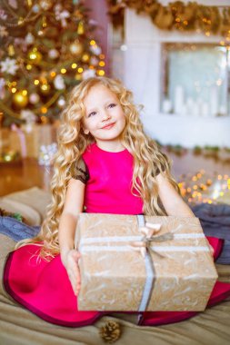 Uzun kıvırcık saçlı sarışın evde hediyeler ve çelenk ve dekore edilmiş bir şömine ile Noel ağacı yakınındaki sevimli küçük kız 