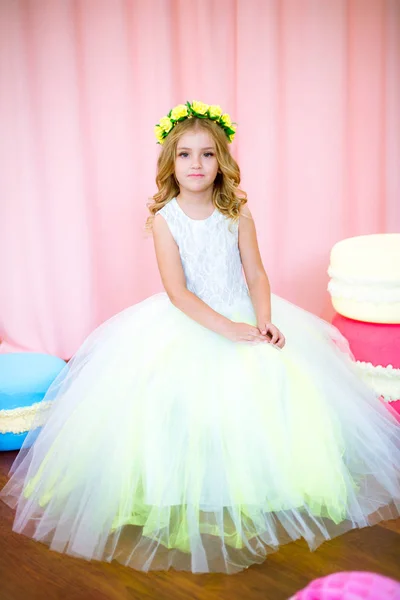 ピンクの背景のスタジオで巨大なお菓子と白い緑豊かなドレスを着たブロンドのカールとかわいい女の子 — ストック写真