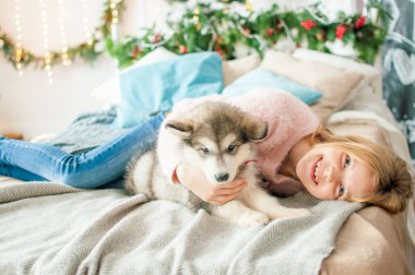 Sarı uzun saçlı sevimli kız Noel için dekore edilmiş bir odada evde küçük Malamute köpek yavrusu ile oynar