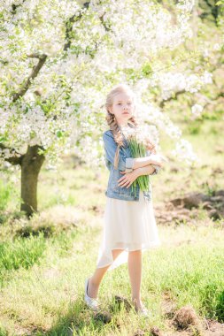 Cute girl with blond hair in a white sundress in spring in a lush garden with daffodils