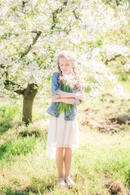 Cute girl with blond hair in a white sundress in spring in a lush garden with daffodils