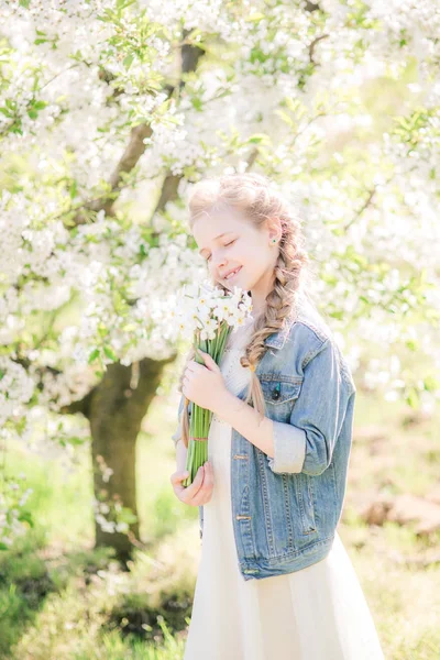 水仙の花と緑豊かな庭園で春に白いサンドレスで金髪のかわいい女の子 — ストック写真