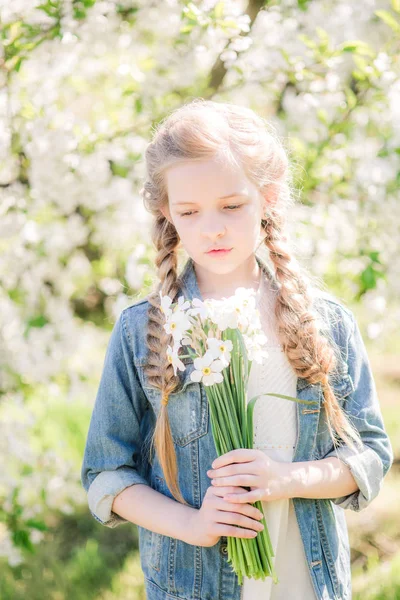 水仙の花と緑豊かな庭園で春に白いサンドレスで金髪のかわいい女の子 — ストック写真