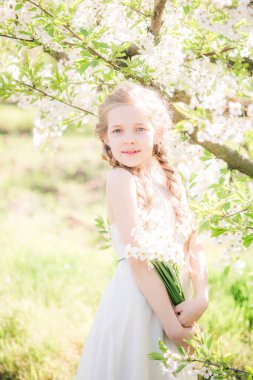 Cute girl with blond hair in a white sundress in spring in a lush garden with daffodils