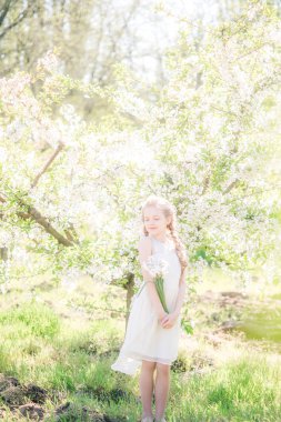 Cute girl with blond hair in a white sundress in spring in a lush garden with daffodils