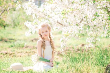 Cute girl with blond hair in a white sundress in spring in a lush garden with daffodils
