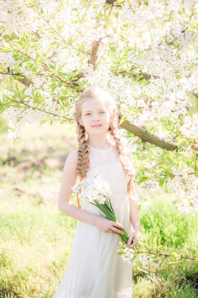 Cute girl with blond hair in a white sundress in spring in a lush garden with daffodils