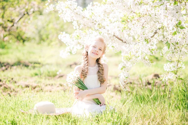 水仙の花と緑豊かな庭園で春に白いサンドレスで金髪のかわいい女の子 — ストック写真