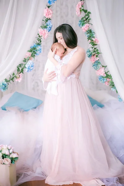 天蓋付きのベッドで春の花の彼女の腕の赤ん坊の私室のドレスで若い母親 — ストック写真