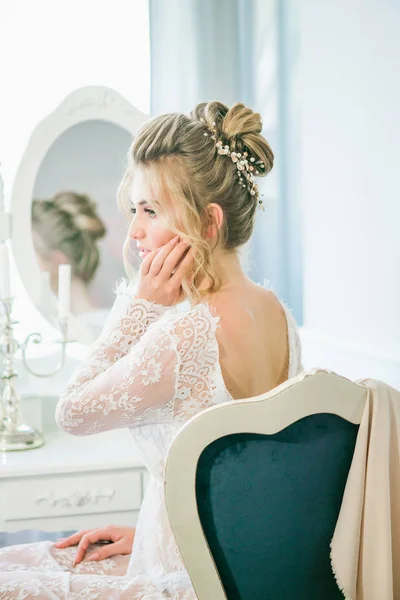 Young cute bride with a beautiful hairdo in the morning at home at the dressing table in white lace negligee