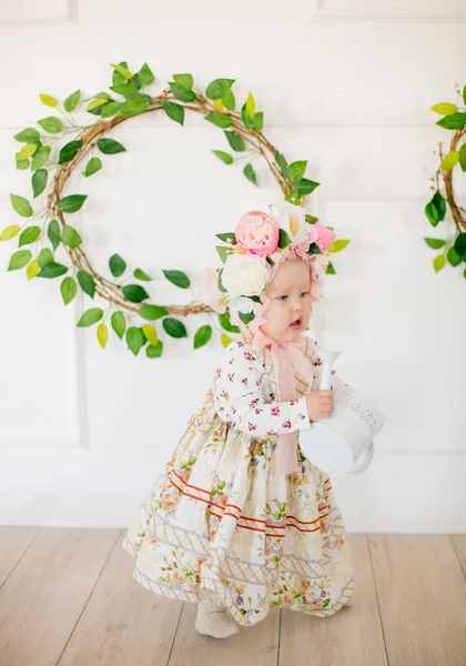 Cute little girl in a dress with a flower print and a hat with flowers in the Easter decorations in the studio. Little girl with easter eggs and flowers in a spring studio