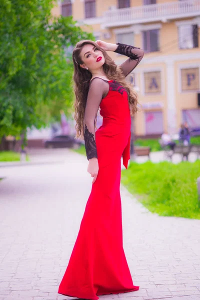 Beautiful young girl with long hair in an evening fashionable red dress with red lipstick on the lips on the street in the public garden