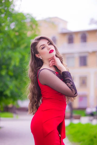 Beautiful young girl with long hair in an evening fashionable red dress with red lipstick on the lips on the street in the public garden