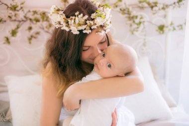Beyaz bir elbiseyle kiraz çiçekleri içinde genç, tatlı bir anne bahar yatak odasında kollarında çiçek açan kiraz dallarıyla bir bebek tutuyor.