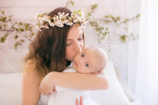 Young cute mom in a wreath of cherry blossoms in a white dress holding a baby in her arms in a spring bedroom with blossoming cherry branches