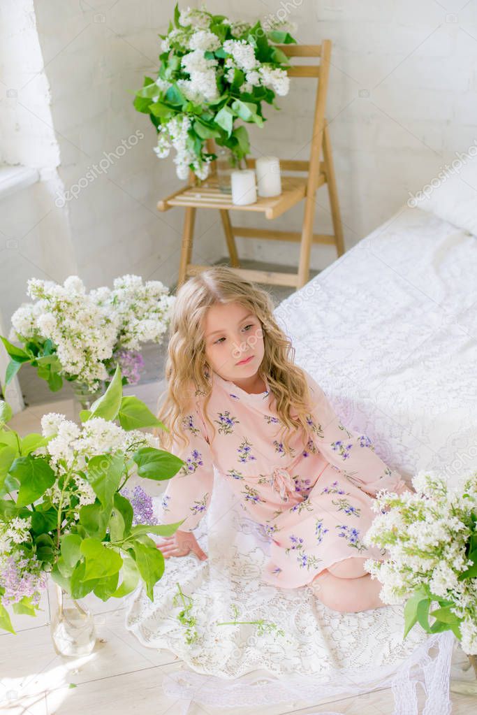 Cute little girl with blond hair in a beautiful dress in a spring studio with lilac flowers