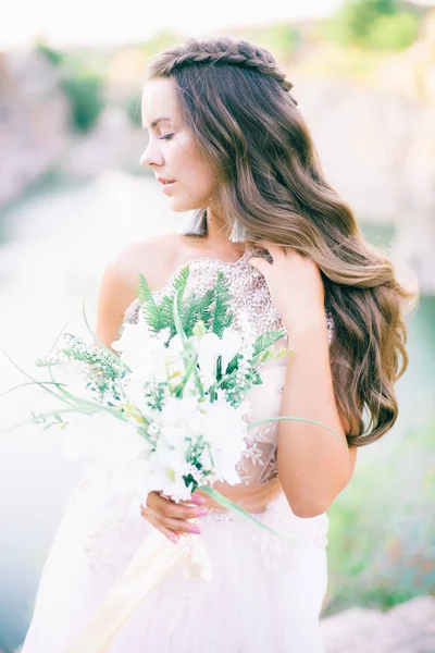 Beautiful young bride with long hair in a wedding dress with a bouquet in the summer on the shore of a beautiful lake