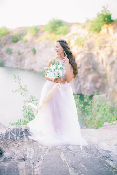 Beautiful young bride with long hair in a wedding dress with a bouquet in the summer on the shore of a beautiful lake