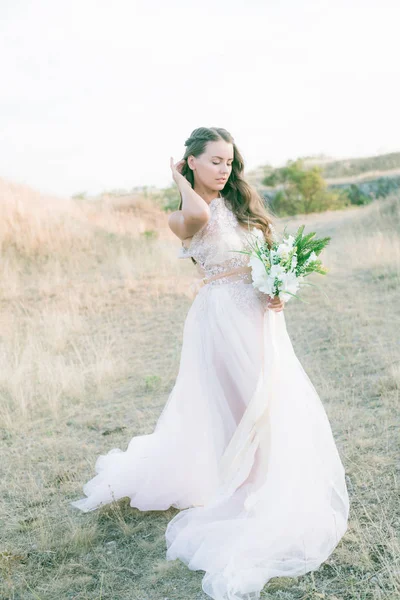 Beautiful young bride with long hair in a wedding dress with a bouquet in the summer in the field