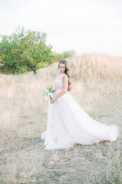 Beautiful young bride with long hair in a wedding dress with a bouquet in the summer in the field