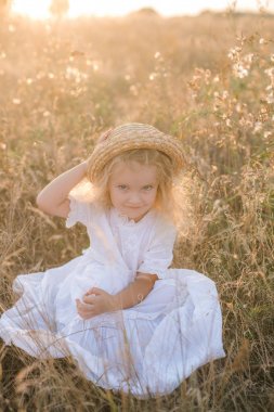 Bir hasır şapka ile beyaz bir elbise ile gün batımında bir yaz alanında sarı saçlı sevimli küçük kız