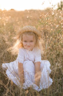 Bir hasır şapka ile beyaz bir elbise ile gün batımında bir yaz alanında sarı saçlı sevimli küçük kız