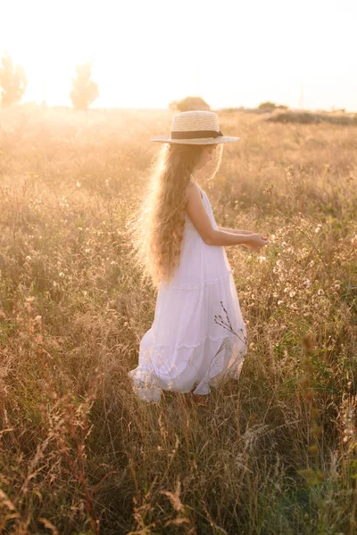 麦わら帽子をかぶった白いドレスを着た夕日の夏のフィールドでブロンドの長い髪を持つかわいい女の子 — ストック写真