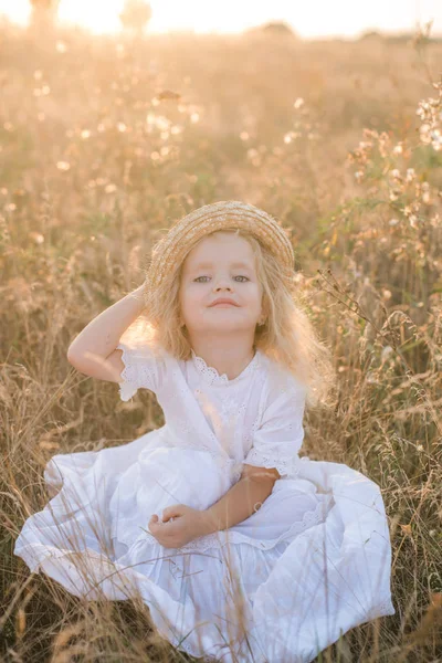 可爱的小女孩与金发在夏日田野在日落与一个白色的礼服与草帽 — 图库照片
