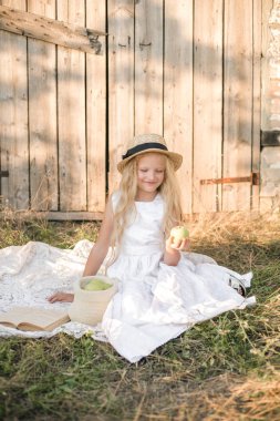 Yeşil elma ile bir hasır şapka ile beyaz bir elbise ile gün batımında bir yaz alanında sarı uzun saçlı sevimli küçük kız