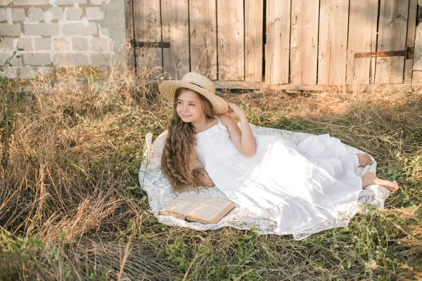 可爱的小女孩与金发长发在夏日田野与白色连衣裙与草帽读书 — 图库照片