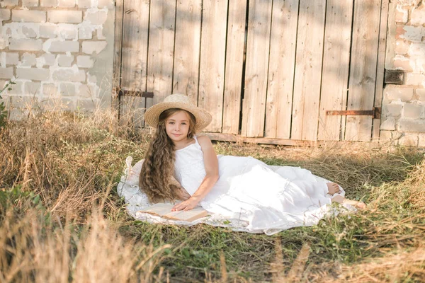 可爱的小女孩与金发长发在夏日田野与白色连衣裙与草帽读书 — 图库照片