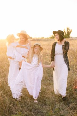 Kızları ile genç bir anne ve kırsal bir alanda yaz gün batımında beyaz elbiseler sarı saçlı bir teyze. Tarlada yaz aylarında aynı aileden birkaç kız