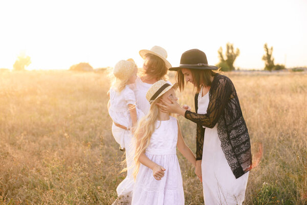 A young mother with her daughters and an aunt with blond hair in white dresses at sunset in the summer in a field of countryside. Several girls from the same family in the summer in the field