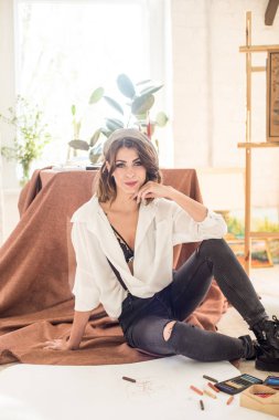 Koyu saçlı çekici kadın sanatçı kağıt ve boya pastel boya ile çevrili bir stüdyoda katta oturur