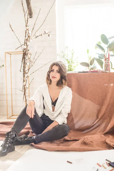 Koyu saçlı çekici kadın sanatçı kağıt ve boya pastel boya ile çevrili bir stüdyoda katta oturur