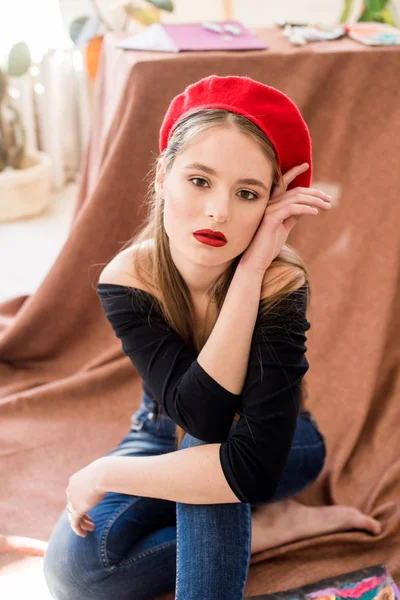長い髪と赤いベレーを持つ魅力的なアーティストの女の子は 紙や塗料のパステルクレヨンに囲まれたスタジオで床に座っています — ストック写真