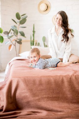 Sade rahat giysiler uzun koyu saçlı güzel genç anne canlı yeşil bitkiler ve doğal malzemelerden yapılmış mobilya ile rahat bir yatak odasında genç oğlu ile oynar