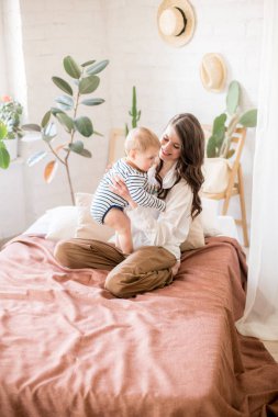 Sade rahat giysiler uzun koyu saçlı güzel genç anne canlı yeşil bitkiler ve doğal malzemelerden yapılmış mobilya ile rahat bir yatak odasında genç oğlu ile oynar