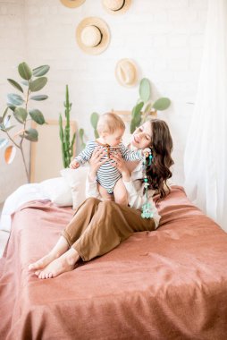 Sade rahat giysiler uzun koyu saçlı güzel genç anne canlı yeşil bitkiler ve doğal malzemelerden yapılmış mobilya ile rahat bir yatak odasında genç oğlu ile oynar
