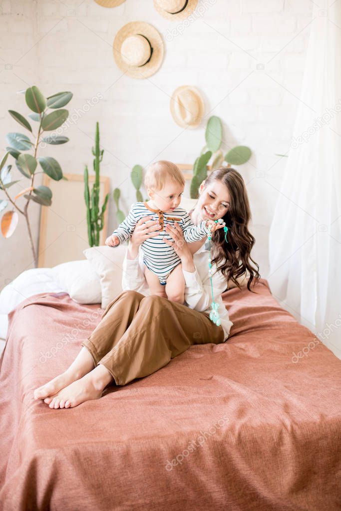 Beautiful young mother with long dark hair in homely comfortable clothes plays with her young son in a cozy bedroom with live green plants and furniture made from natural materials