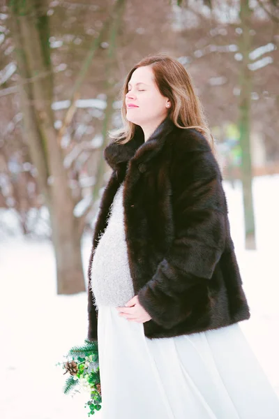 公園の雪の上で冬に花束を持つミンクのコートと長いドレスを着た若い妊娠中の女性 — ストック写真