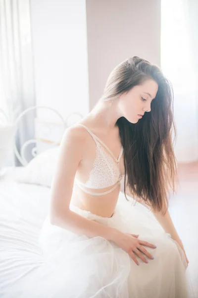 年轻美丽的新娘与黑色长发早晨在家在白色的床上 在白色蕾丝紧身衣和礼服 — 图库照片