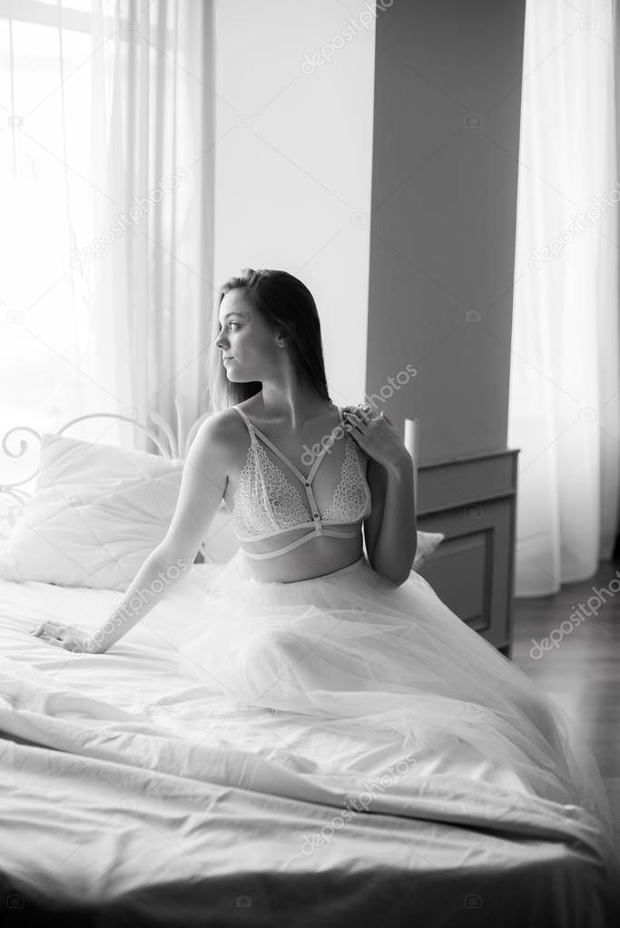 Young beautiful bride with dark long hair morning at home on a white bed in a white lace bustier and dress