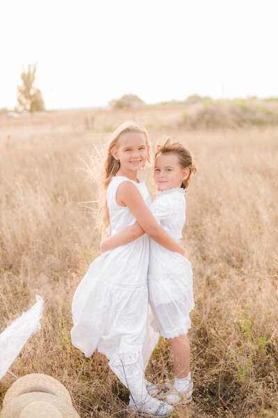Милые маленькие девочки-сёстры с светлыми волосами на летнем поле на закате в белых платьях в соломенной шляпе
